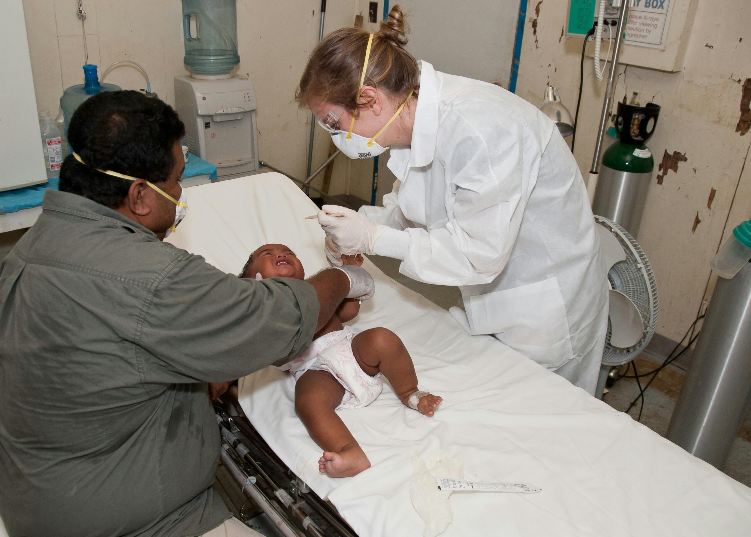 Doctor in protective gear treating a baby on a hospital bed with a parent present.